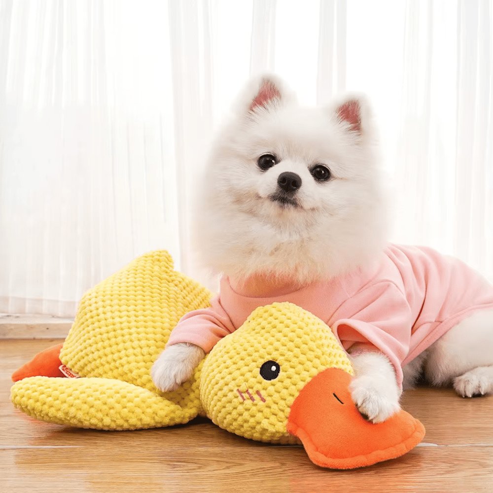 Yellow plush duck dog toy with orange bill and feet, textured fabric, lying on a light surface (view 2)
